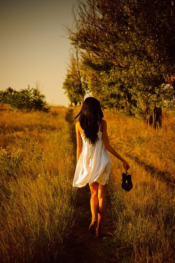 The barefoot girl in white dress with shoes in hand is on the field. Rear view